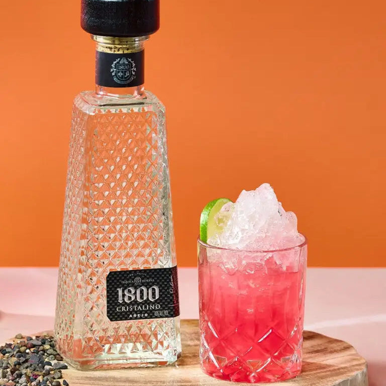 Load image into Gallery viewer, Bottle of 1800 Cristalino Añejo Jose Cuervo Tequila with a pink cocktail on a wooden board against an orange background