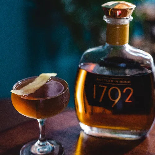 Bottle of 1792 bourbon and a cocktail with a lemon twist on a wooden surface.