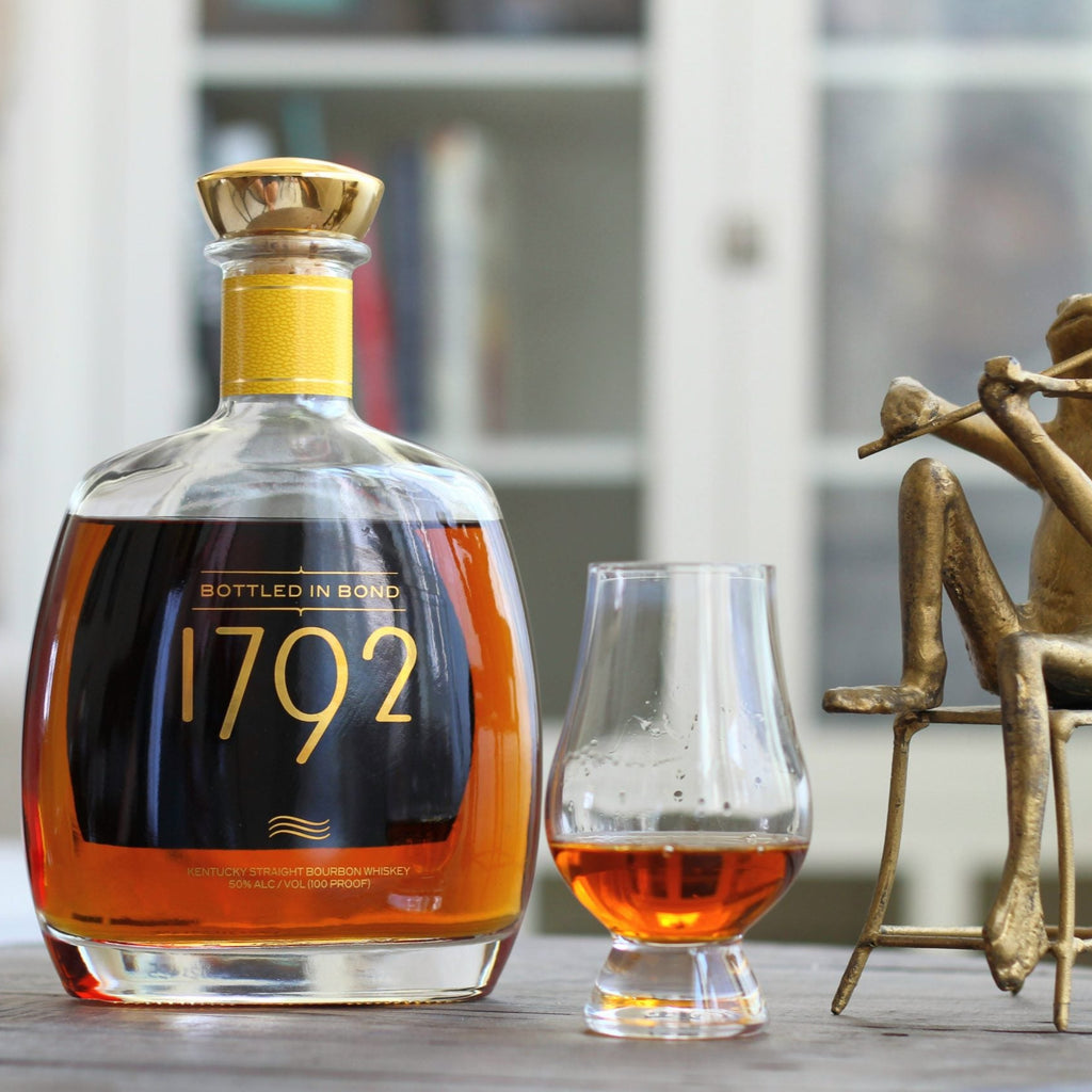 Bottle of 1792 bourbon with a glass of whiskey on a table, accompanied by a decorative figure.