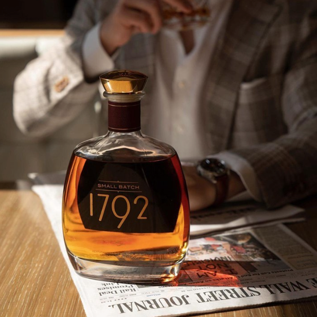 Load image into Gallery viewer, A bottle of 1792 Small Batch bourbon whiskey sits on a newspaper on a wooden table, with a man in a plaid blazer holding a glass of whiskey in the background.
