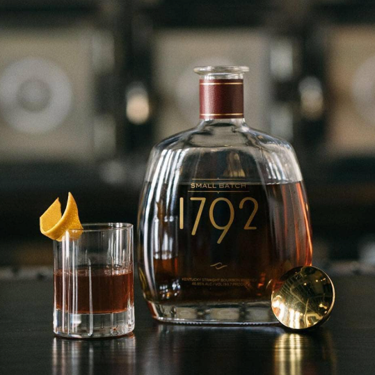 Load image into Gallery viewer, A clear glass bottle of Small Batch 1792 bourbon whiskey stands on a dark bar counter beside a matching glass filled with amber liquor and garnished with a lemon twist, with blurred bottles in the background.