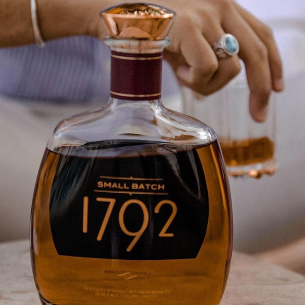 Load image into Gallery viewer, A bottle of 1792 Small Batch bourbon whiskey on a tabletop, with a person holding a glass in the softly blurred background.