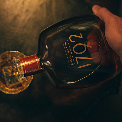 Load image into Gallery viewer, A hand holding a dark glass bottle of 1792 Small Batch bourbon whiskey, pouring a clear amber liquid into a cut glass on a dark wooden surface.