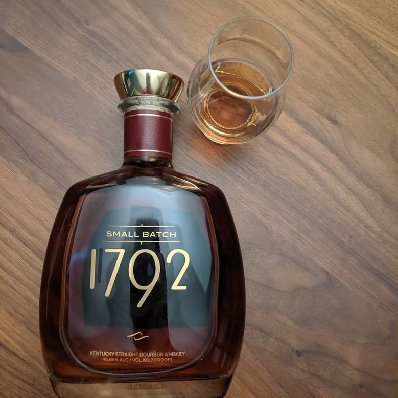 Load image into Gallery viewer, A bottle of 1792 Small Batch Kentucky Straight Bourbon Whiskey with a gold cap, shown next to a glass of the whiskey on a dark wood tabletop.