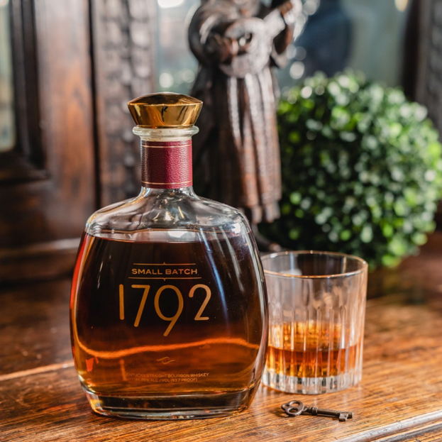 Load image into Gallery viewer, A bottle of 1792 Small Batch bourbon whiskey with a gold cap and a matching glass of amber liquor on a rustic wooden table, with a small key and blurred decorative statue in the background.
