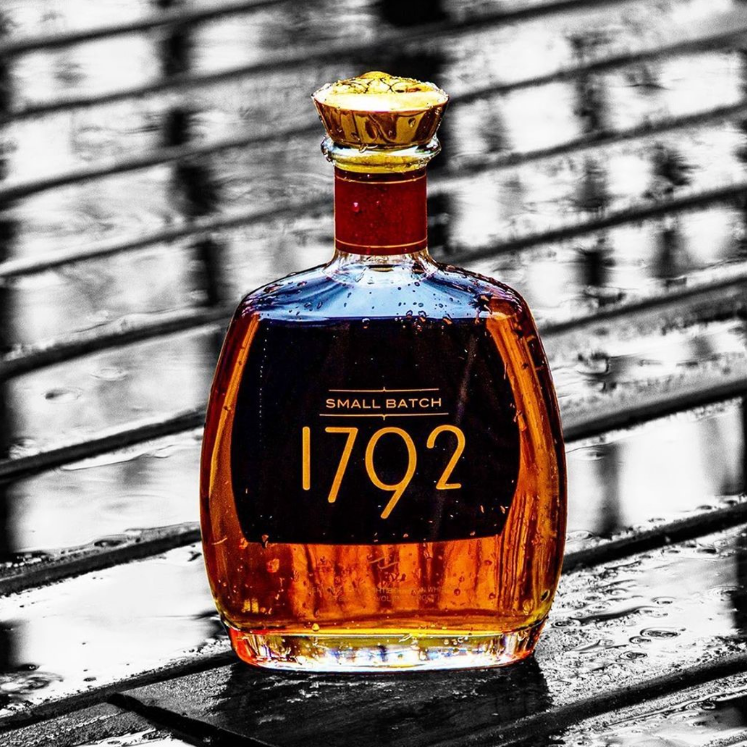 Load image into Gallery viewer, A bottle of 1792 Small Batch bourbon whiskey with a gold cap, photographed on a wet dark wooden surface with a blurred black-and-white background.