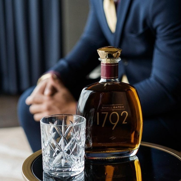 Load image into Gallery viewer, A bottle of Small Batch 1792 whiskey and a clear cut-glass tumbler resting on a round black tray with a gold rim, with a man in a navy suit blurred in the background.