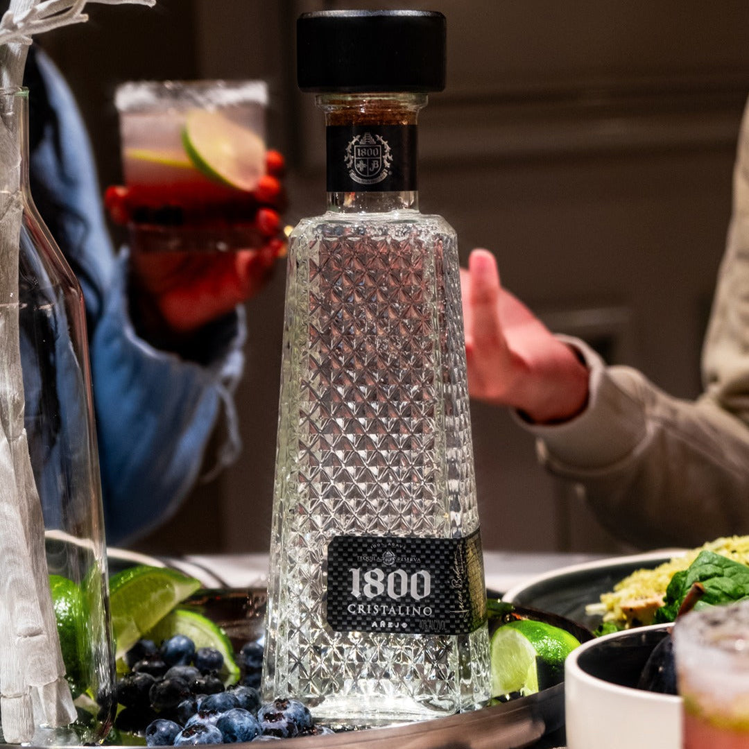 Load image into Gallery viewer, 1800 Cristalino Añejo Jose Cuervo Tequila bottle on a table with people and food in the background