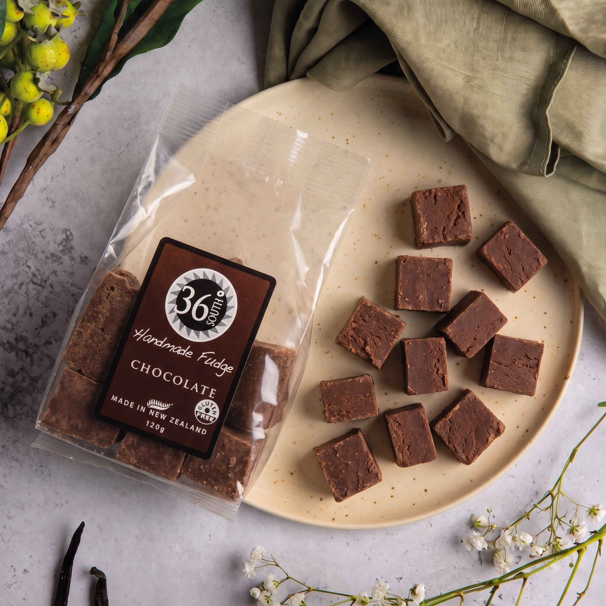 Load image into Gallery viewer, A clear bag of 36 Degrees South Handmade Fudge Chocolate squares sits beside a beige plate holding several chocolate fudge pieces, styled with a folded olive cloth and small floral accents on a light grey surface.