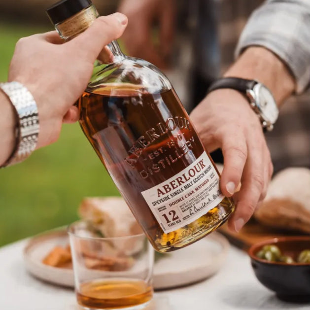 Load image into Gallery viewer, Person holding a bottle of Aberlour 12yo whisky with a glass on a casual outdoor setting