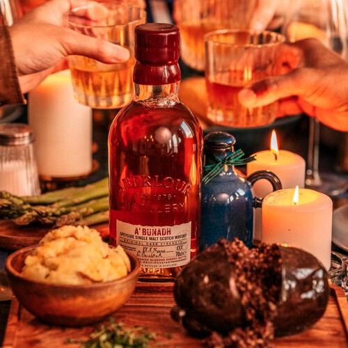 A bottle of Aberlour Abundah whisky on a table with candles and food, people clinking glasses in the background