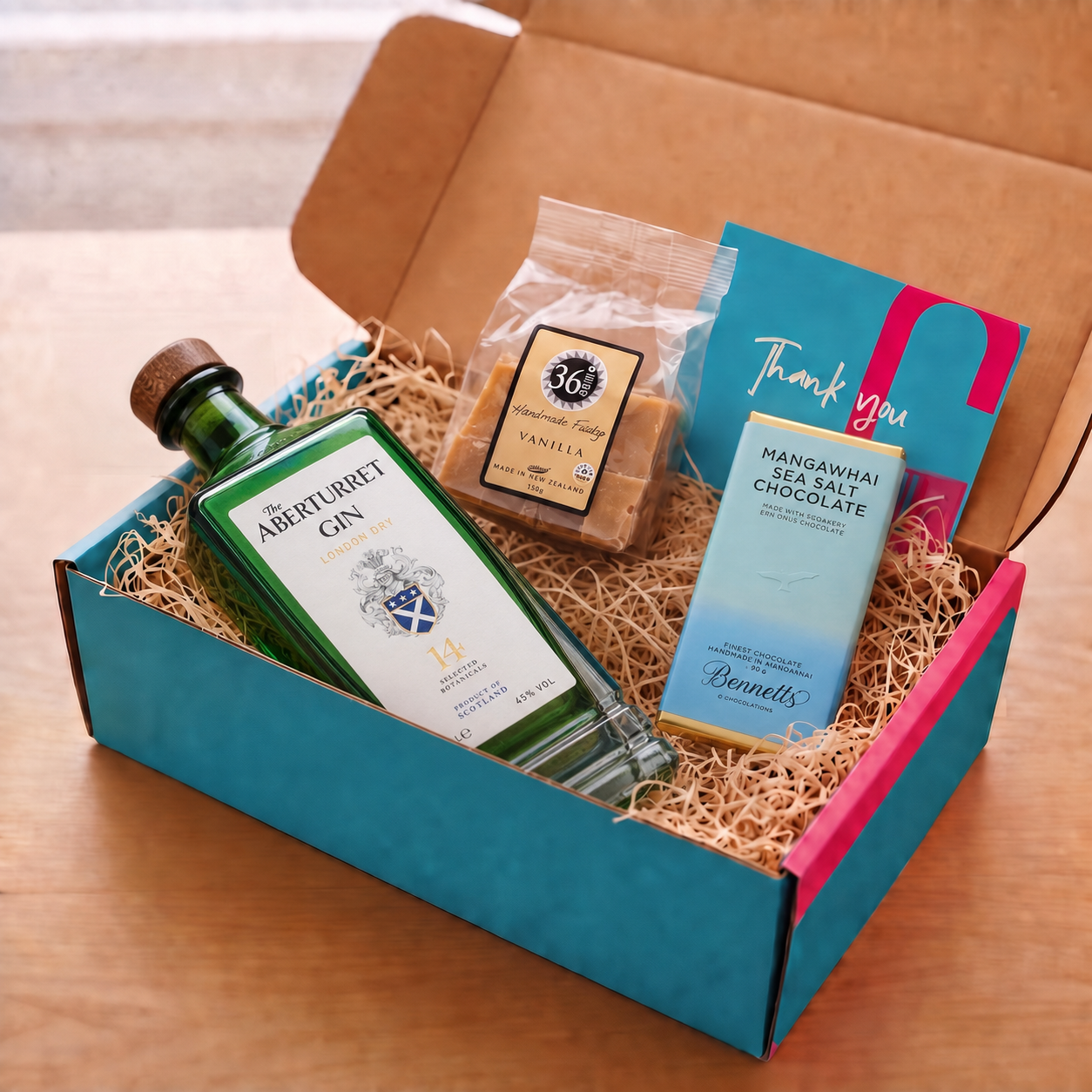 Load image into Gallery viewer, A curated gift box set on a wooden table, featuring a bottle of Aberturbet Gin, a bag of 36 Degrees Vanilla fudge, a bar of Bennett's Mangawhai Sea Salt Chocolate, and a blue 'Thank you' card, all arranged in a turquoise and pink COVE 27 box with wood-wool.
