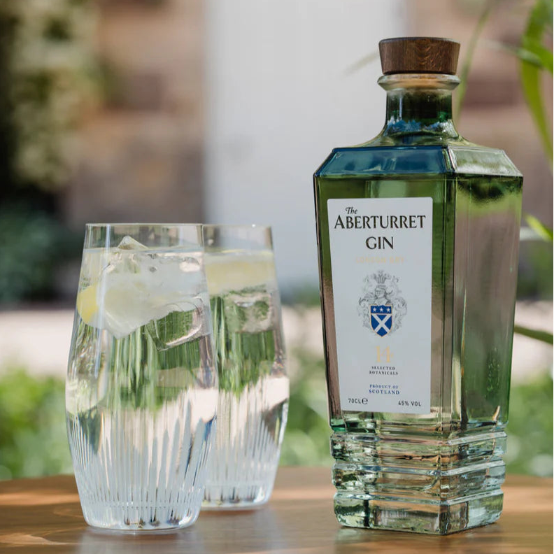 Load image into Gallery viewer, A bottle of Aberturret London Dry Gin next to two clear glasses with ice and lemon on a wooden table, set against a blurred outdoor garden background.