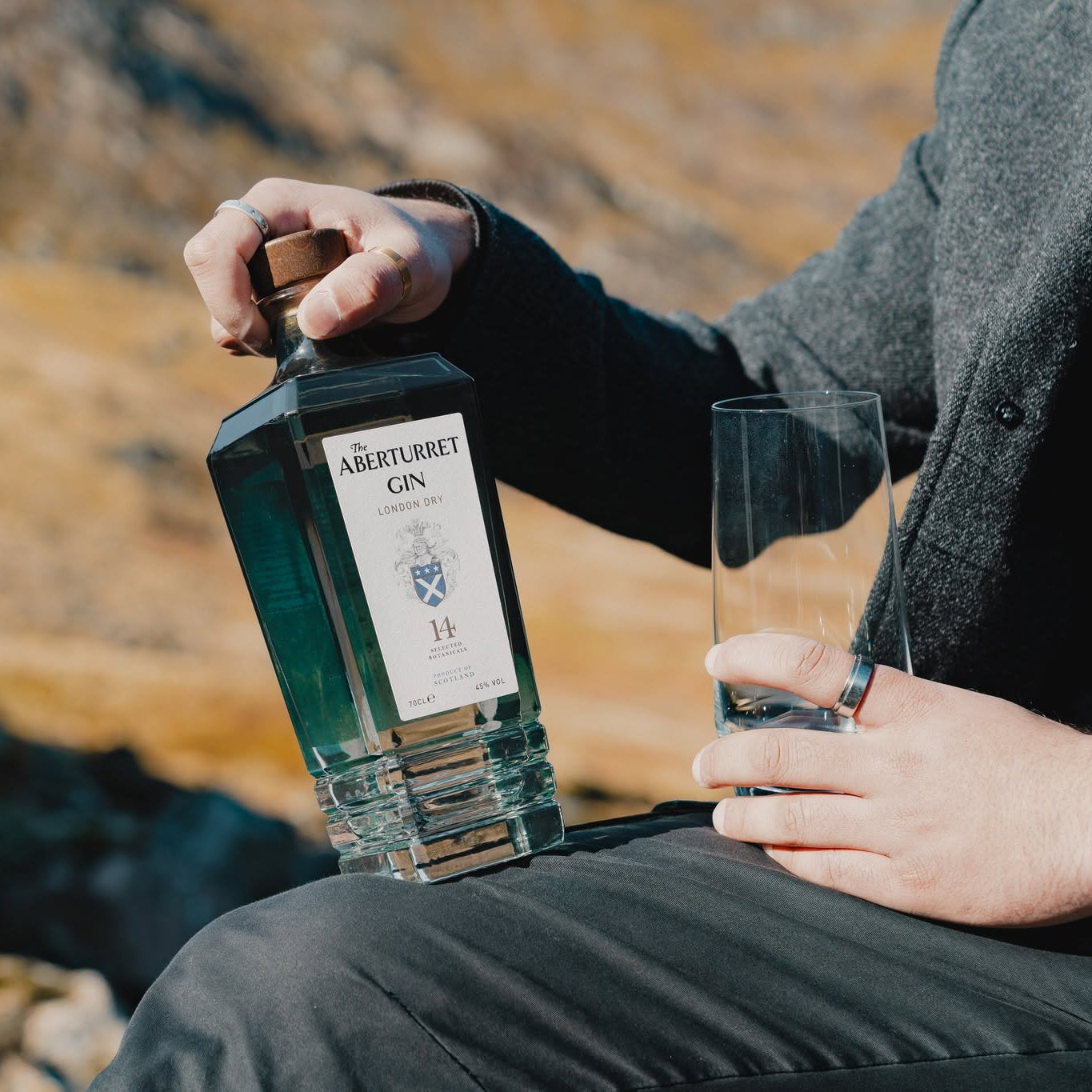 Load image into Gallery viewer, A person outdoors holds a bottle of Aberturret London Dry Gin next to a clear glass, with a blurred mountain landscape in the background.