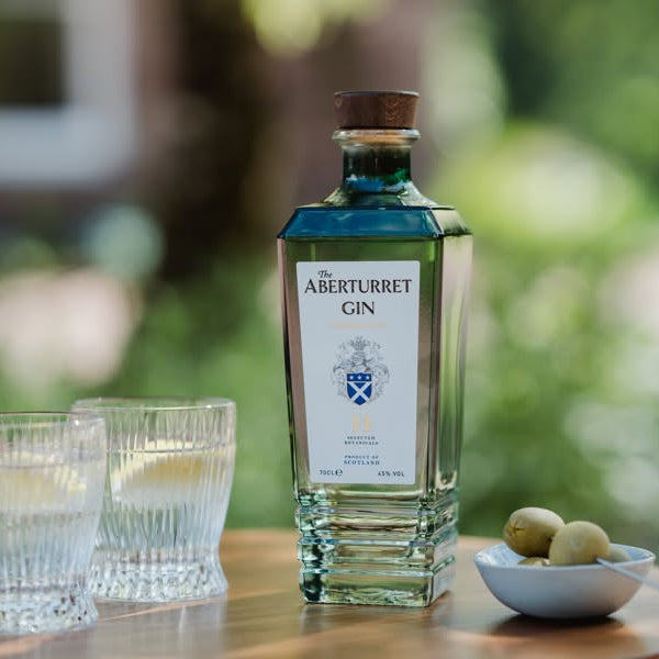 Load image into Gallery viewer, A bottle of Aberturret London Dry Gin with two clear glasses and a small bowl of olives on a wooden table, set against a blurred green outdoor background.