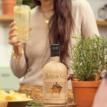 Load image into Gallery viewer, Woman holding a glass of juice next to a bottle of Bathtub Gin in a kitchen setting.