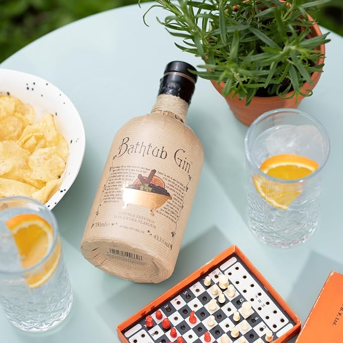 Load image into Gallery viewer, A bottle of Bathtub Gin lies on a pale blue table surrounded by snacks and drinks, including chips, glasses with orange slices, and a potted green plant. An orange game board is also visible.