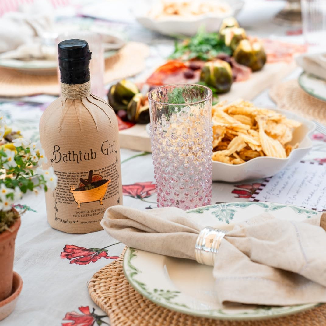 Load image into Gallery viewer, A styled dining table setting with a beige linen bottle labeled 'Bathtub Gin' as the main product. The scene includes a pink textured drinking glass, a white bowl of chips, plates with folded beige napkins, woven placemats, and a floral tablecloth in a bright, softly lit indoor environment.