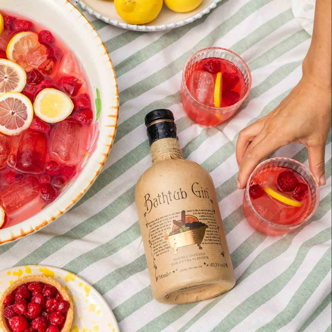 Load image into Gallery viewer, A bottle of Bathub Gin lies on a green-and-white striped picnic blanket surrounded by a large bowl of pink berry and lemon iced drink, two clear glasses with pink drinks and lemon slices, and a hand reaching toward one glass. Plates with cherries and lemons are also visible.