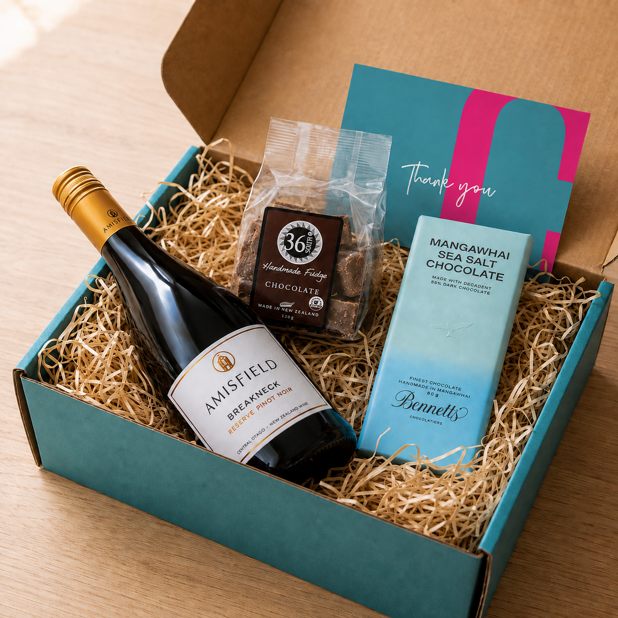 Load image into Gallery viewer, A curated gift box containing an Amisfield Breakneck Reserve Pinot Noir red wine bottle, a bag of 36 Degrees Handmade Fudge Chocolate, a light blue bar of Bennett's Mangawhai Sea Salt Chocolate, and a teal thank you card, arranged in natural wood-wool inside an open COVE 27 teal box on a wooden table.