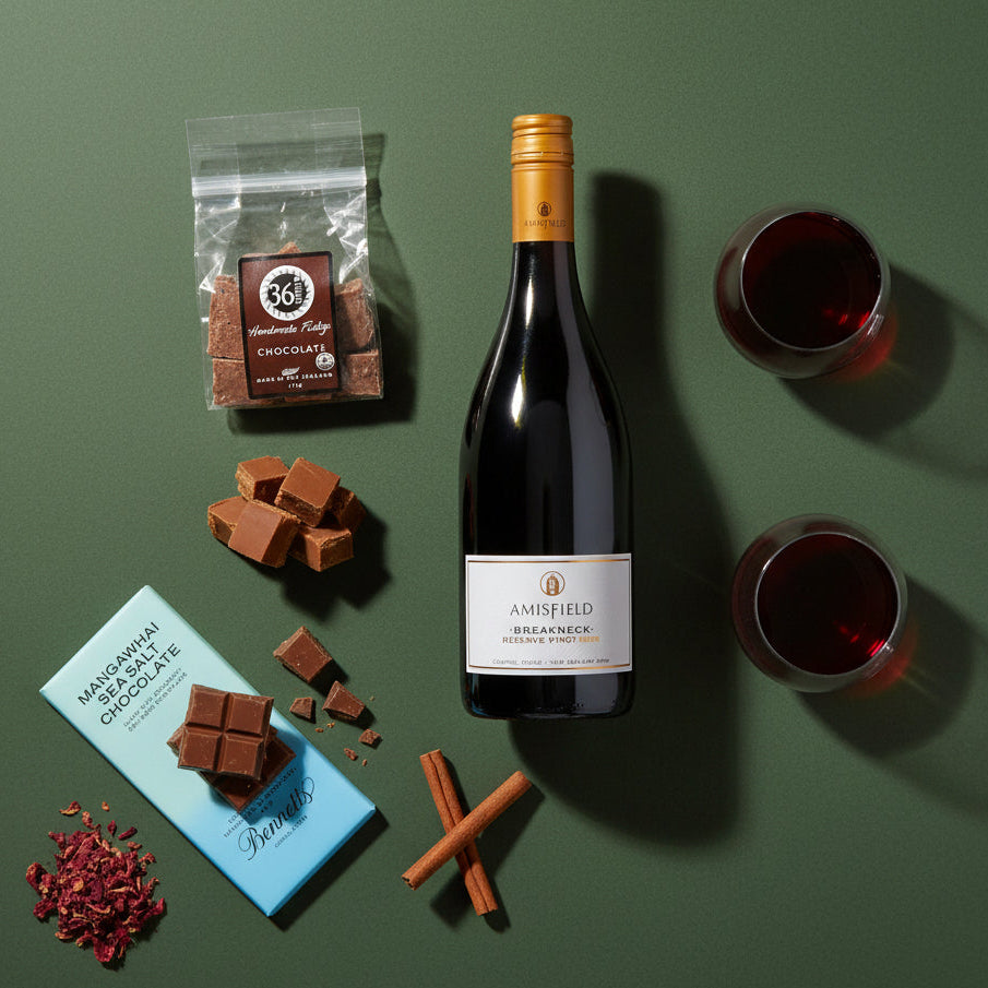 Load image into Gallery viewer, A bottle of Amisfield Breaknech Reserve Pinot Noir red wine is arranged on a dark green background with two filled wine glasses, a bag of 36 Degrees chocolate fudge, loose chocolate pieces, cinnamon sticks, and a blue Bennett's chocolate bar.