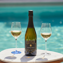 Load image into Gallery viewer, Amisfield Methode Traditionelle Brut sparkling wine bottle next to two half-full champagne flutes on a table with a swimming pool in the background