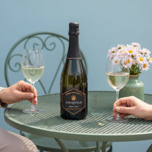 Load image into Gallery viewer, A bottle of Amisfield Methode Traditionelle Brut sparkling wine next to two champagne flutes being held by a woman and man on an outdoor table with pink flowers behind.