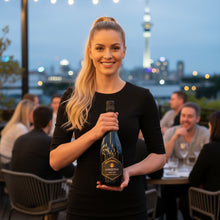 Load image into Gallery viewer, A woman waitress holding a bottle of Amisfield Methode Traditionelle Brut sparkling wine in the outside area of an Auckland restaurant with the Sky Tower in the background.