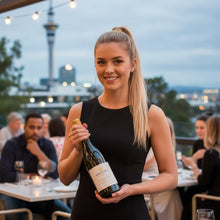 Load image into Gallery viewer, Woman holding a Amisfield Central Otago Sauvignon Blanc wine bottle in an outdoor setting with people and lights in the background