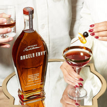 Load image into Gallery viewer, Bottle of Angels Envy bourbon with a person holding a glass and another person garnishing a cocktail.