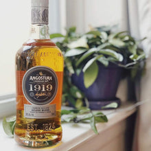 Load image into Gallery viewer, Angostura 1919 rum bottle on a windowsill with plants in the background