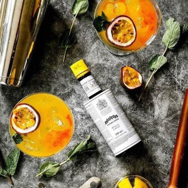 Cocktails with passion fruit, a bottle of Angostura Aromatic Bitters, and bar tools on a dark surface.