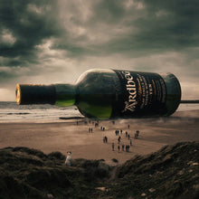 Load image into Gallery viewer, Large Ardbeg 10 year old whisky bottle on a beach with people in the distance.