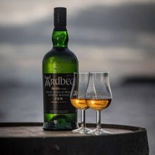Load image into Gallery viewer, Ardbeg 10 year old whisky bottle with two glasses on a barrel against a blurred coastal background