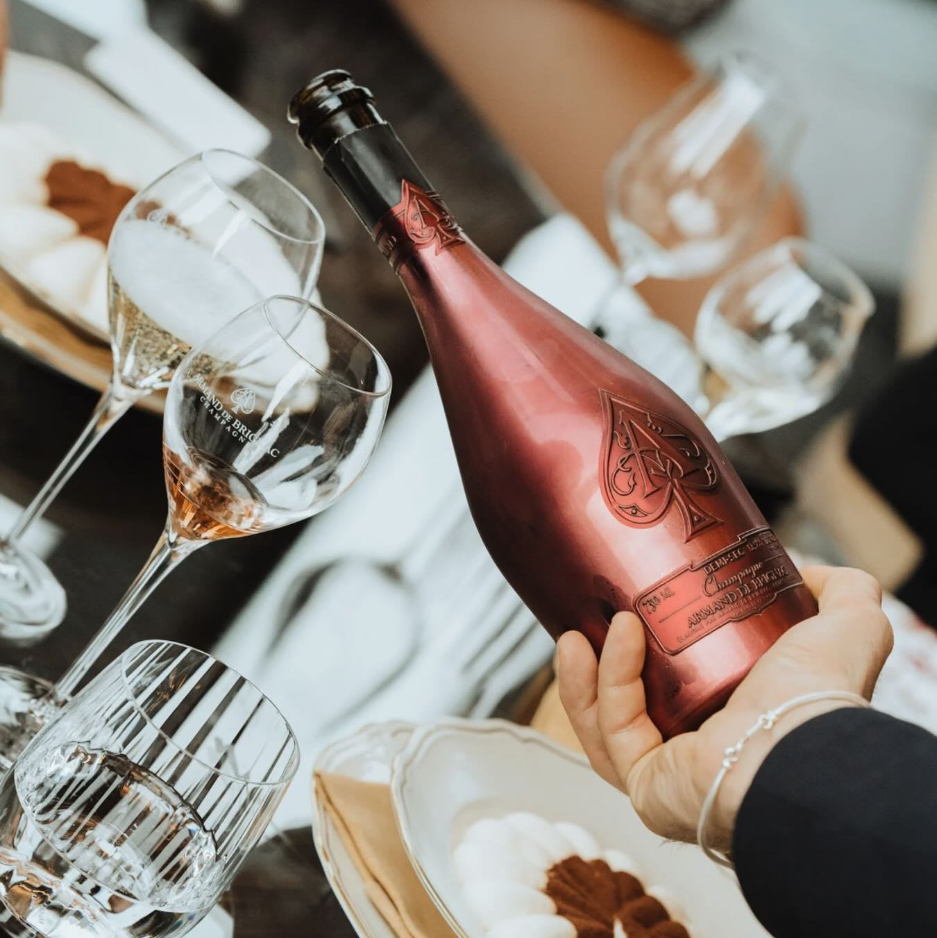Load image into Gallery viewer, A hand holding a pink bottle of Armand de Brignac Ace of Spades Rosé Champagne with a metallic rose gold foil label, surrounded by wine glasses and plates on a dining table.