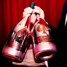 Load image into Gallery viewer, Two pink bottles of champagne being held against a red curtain background.