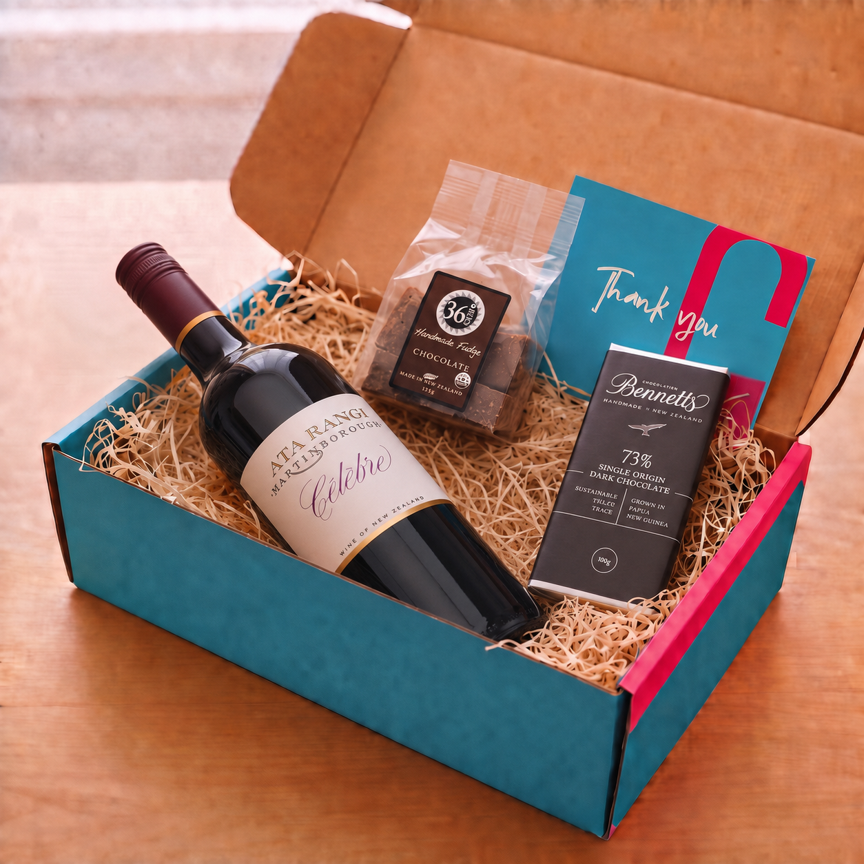 Load image into Gallery viewer, A curated gift box set on a wooden table, featuring a bottle of Ata Rangi Celebre Red wine, a bag of 36 Degrees chocolate fudge, a bar of Bennetts dark chocolate, and a blue and pink thank you card inside a teal and pink box.