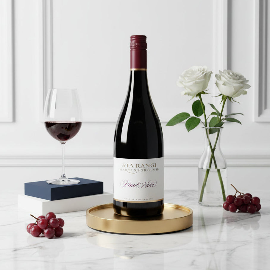 Load image into Gallery viewer, A bottle of Ata Rangi Pinot Noir red wine stands on a round gold tray on a white marble surface, styled with a glass of red wine, white roses in a vase, and clusters of red grapes against a white panelled wall background.