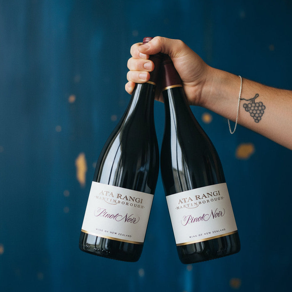 Load image into Gallery viewer, A hand holding two bottles of Ata Rangi Marlborough Pinot Noir wine against a dark blue textured background with gold speckles.