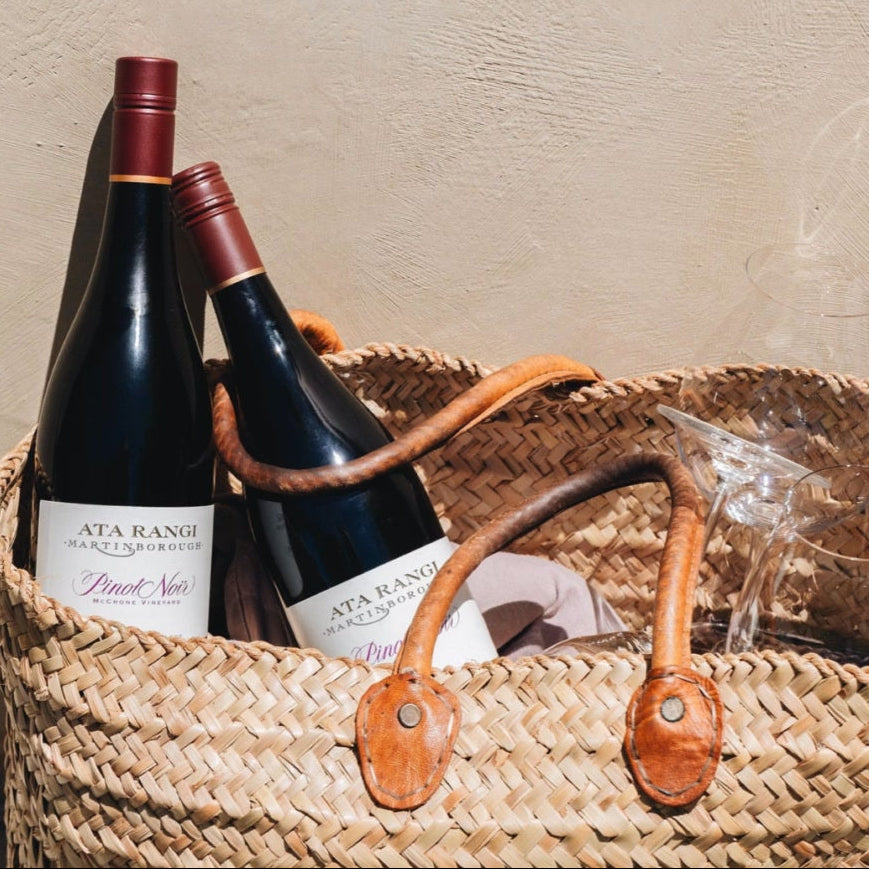 Load image into Gallery viewer, A woven straw tote bag with tan leather handles sits on a beige wall, holding two Ata Rangi Pinot Noir wine bottles and a clear glass. A small framed botanical print hangs on the wall above.