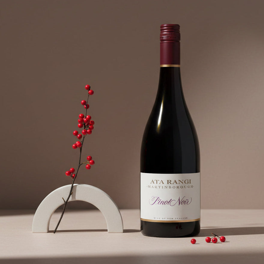 Load image into Gallery viewer, A bottle of Ata Rangi Pinot Noir wine stands on a beige surface against a warm taupe background, styled with a small white arch holder and red berries.