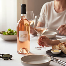 Load image into Gallery viewer, Person holding a glass of rose wine with a bottle of BY.OTT Provence Rose Magnum at a dining table.
