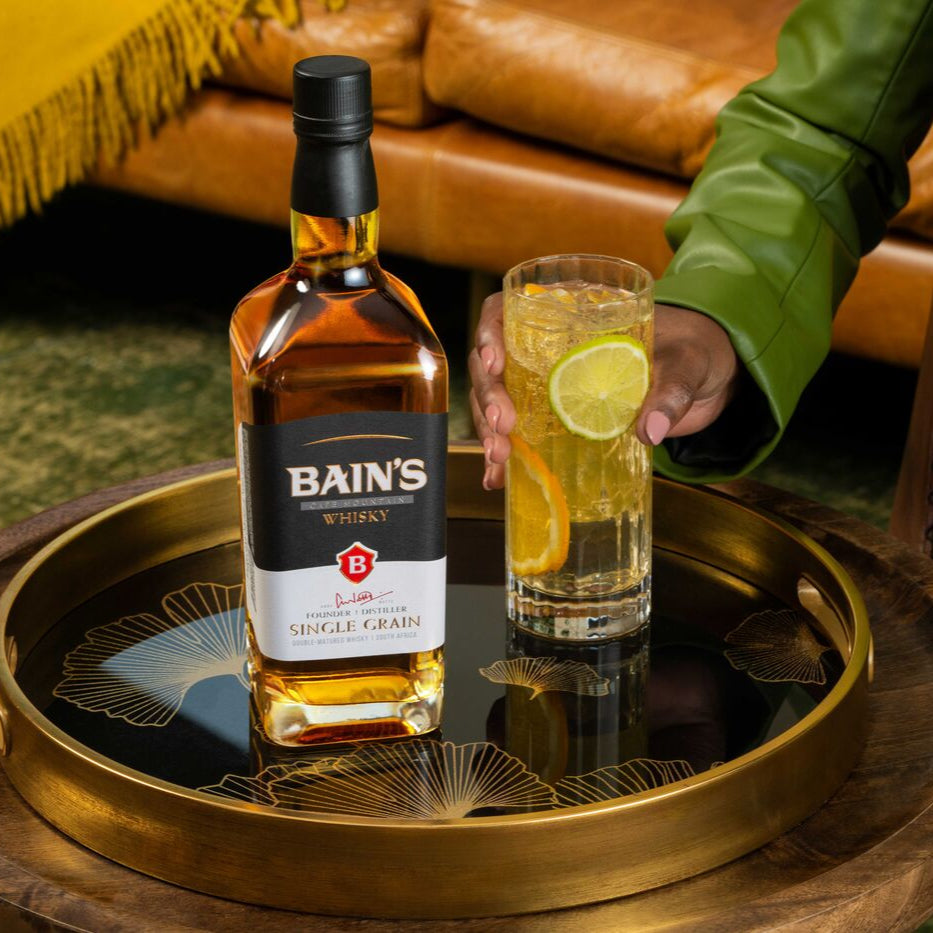 Bain's Single Grain Whisky bottle on a wooden tray with a glass of lime and orange juice, person in green jacket in background.