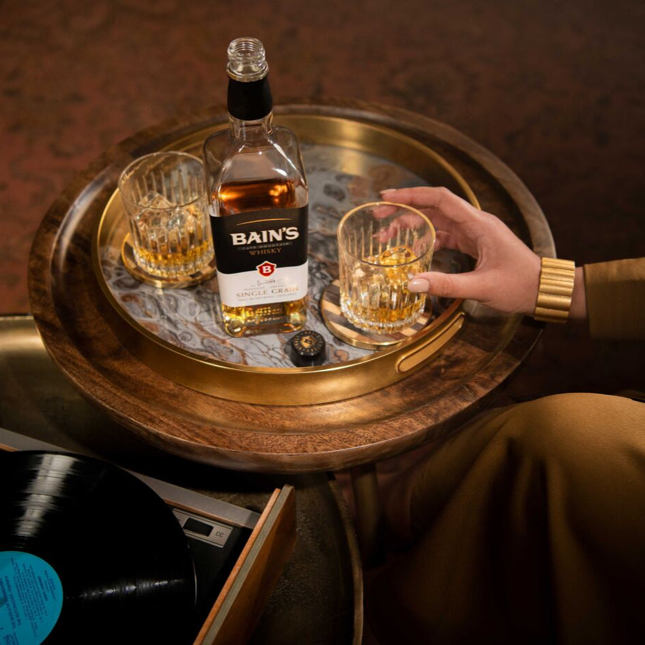 Bottle of Bains Grain whiskey with two glasses on a wooden tray, with a person holding one glass.