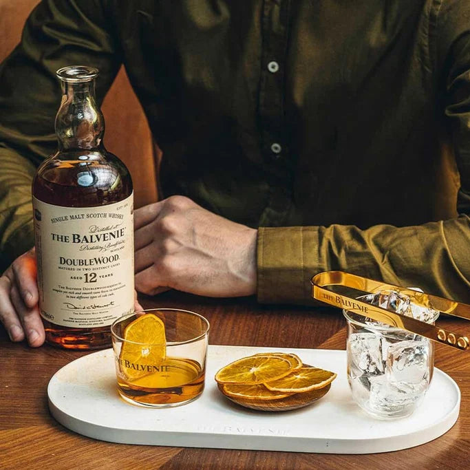 Load image into Gallery viewer, A bottle of The Balvenie Doublewood 12 Year Old Single Malt Scotch Whisky is displayed on a wooden table next to two glasses with orange slices, ice, and tongs, with a person's hands and torso visible in the background.