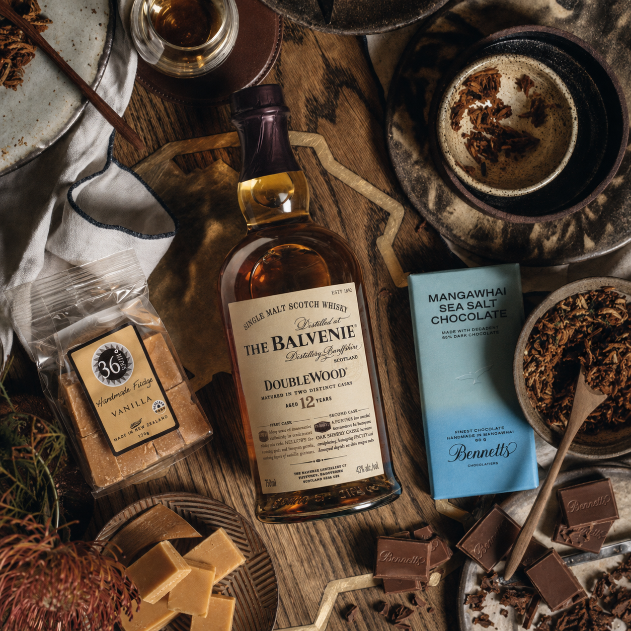 Load image into Gallery viewer, A styled flat-lay featuring a bottle of The Balvenie Doublewood 12YO Single Malt Scotch Whisky alongside a blue bar of Bennetts Mangawhai Sea Salt Chocolate, surrounded by chocolate pieces, sea salt, and dried botanicals on a rustic wooden table.