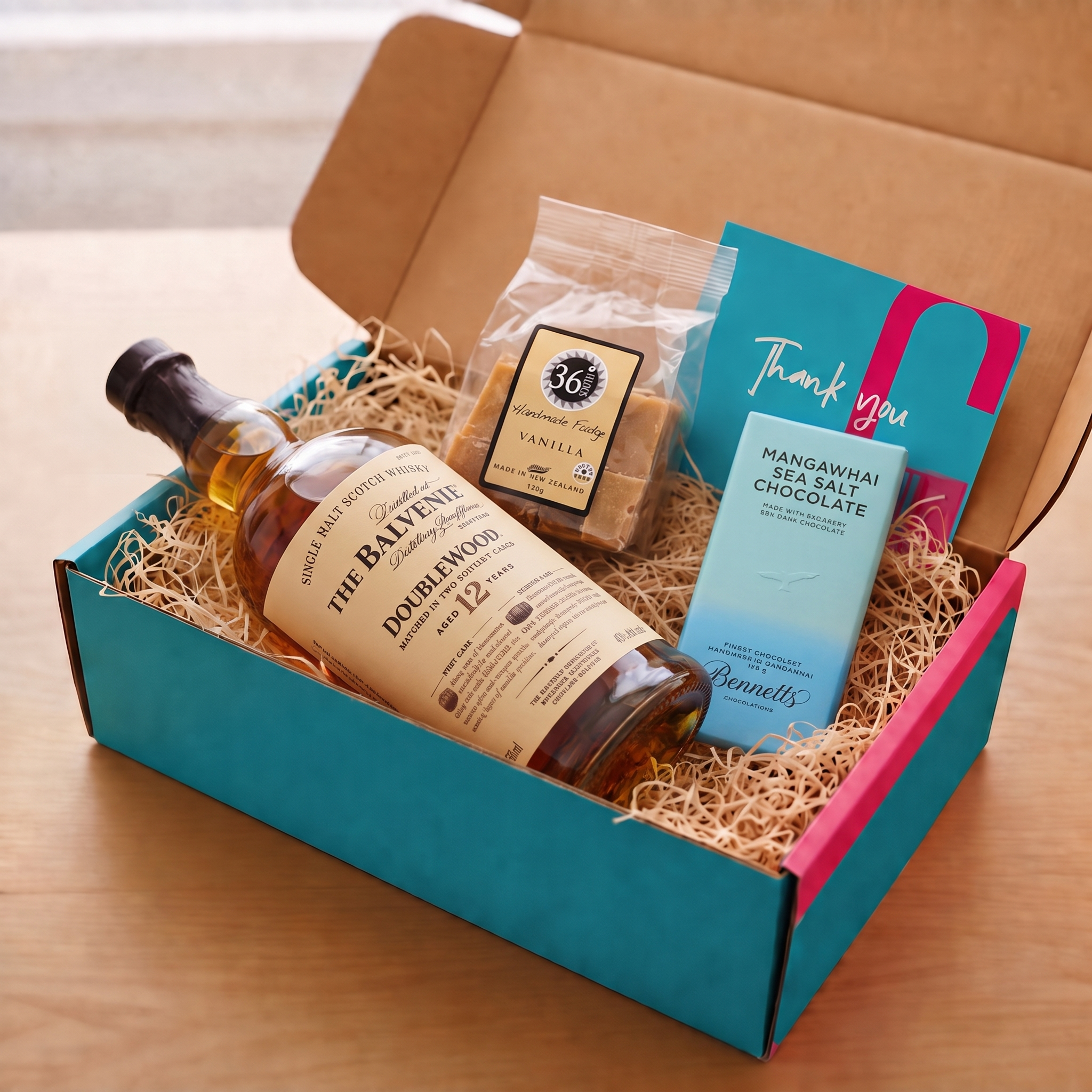 Load image into Gallery viewer, An open gift box filled with cwood-wool on a wooden surface, containing a bottle of The Balvenie Doublewood 12yo Single Malt Scotch, a bag of 36 Degrees Vanilla fudge, a blue Bennetts Mangawhai Sea Salt Chocolate bar, and a pink and blue Thank You card.