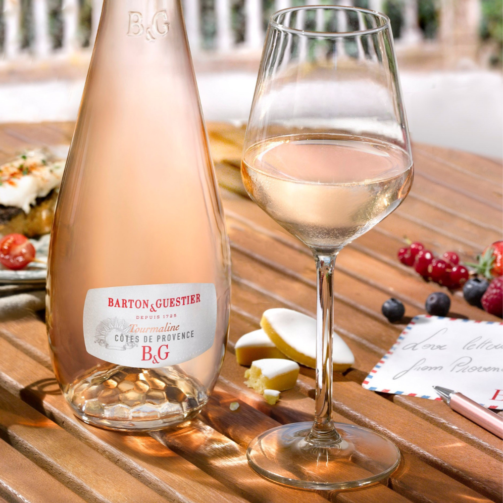Bottle and glass of Barton & Guestier rosé wine on a wooden table with food and fruit.