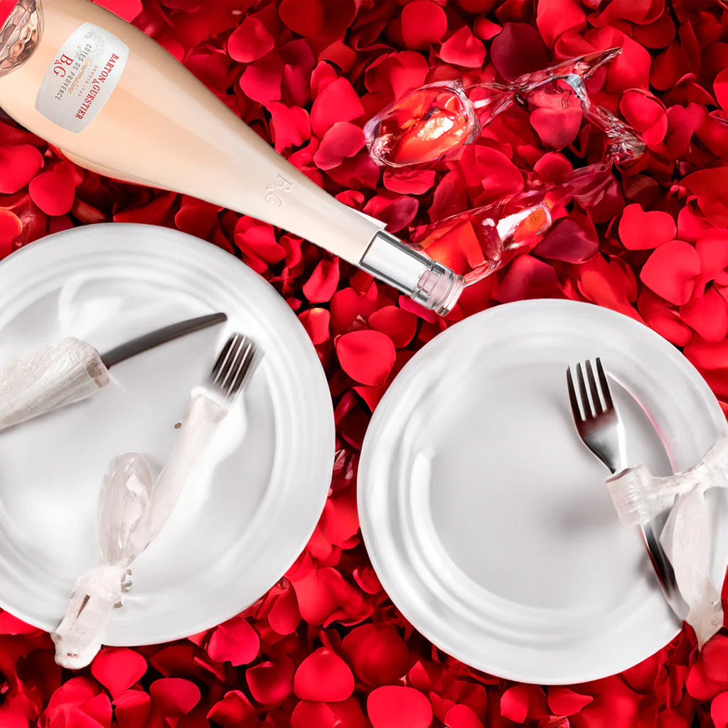 Bottle of champagne and glasses on a bed of red rose petals with white plates and cutlery.