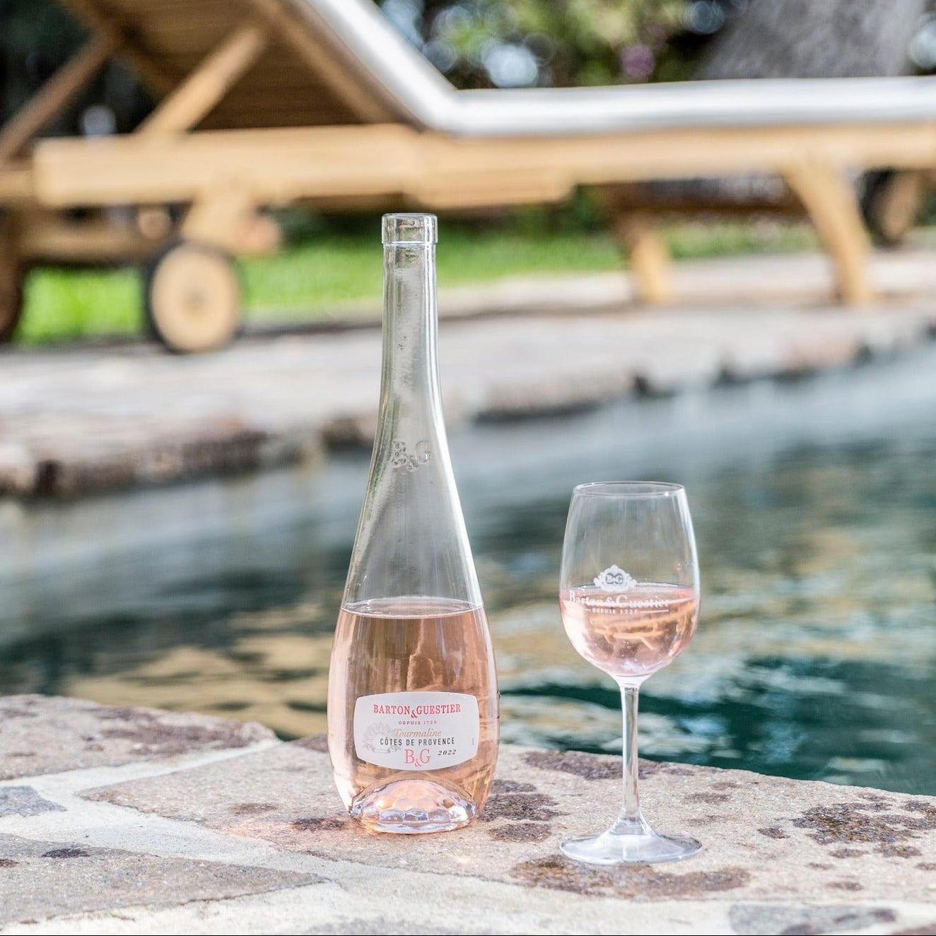 Load image into Gallery viewer, Bottle and glass of rosé wine by a poolside with lounge chair and garden.